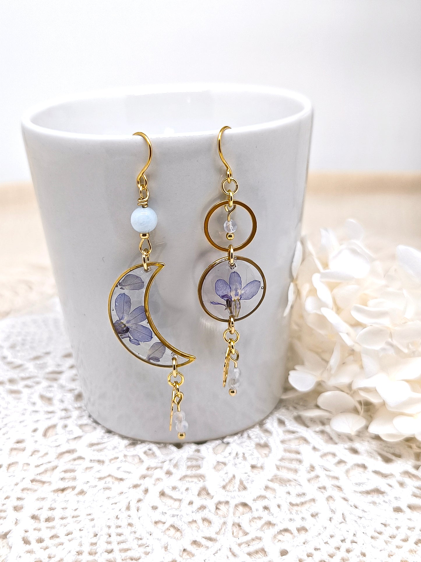 Boucles d'oreilles asymétriques en forme de lune et de soleil contenant une fleur séchée et des pierres fines.