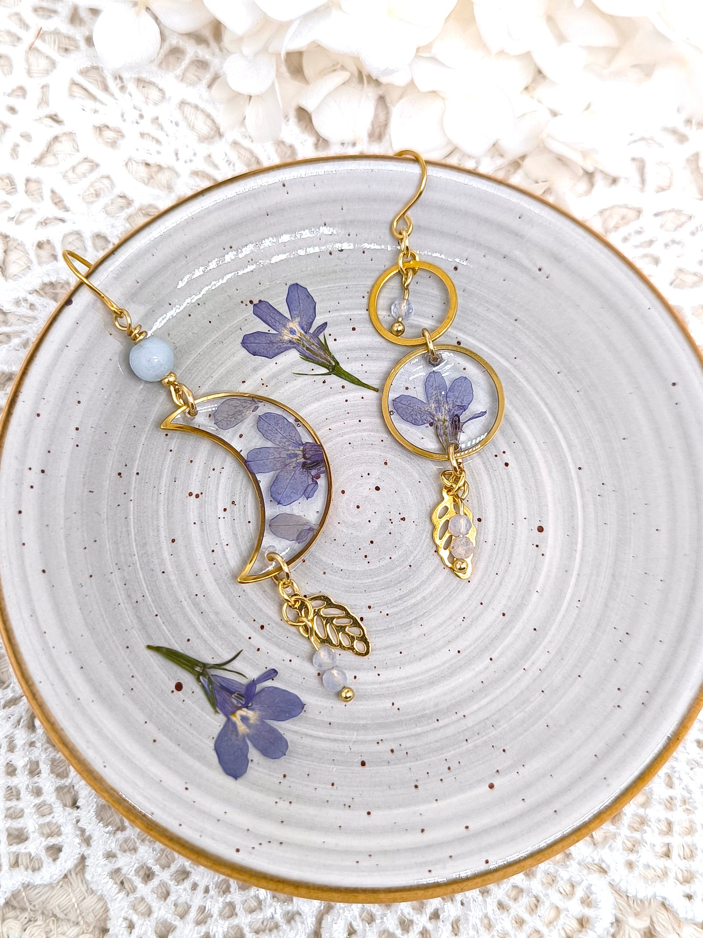 Boucles d'oreilles asymétriques en forme de lune et de soleil contenant une fleur séchée et des pierres fines.
