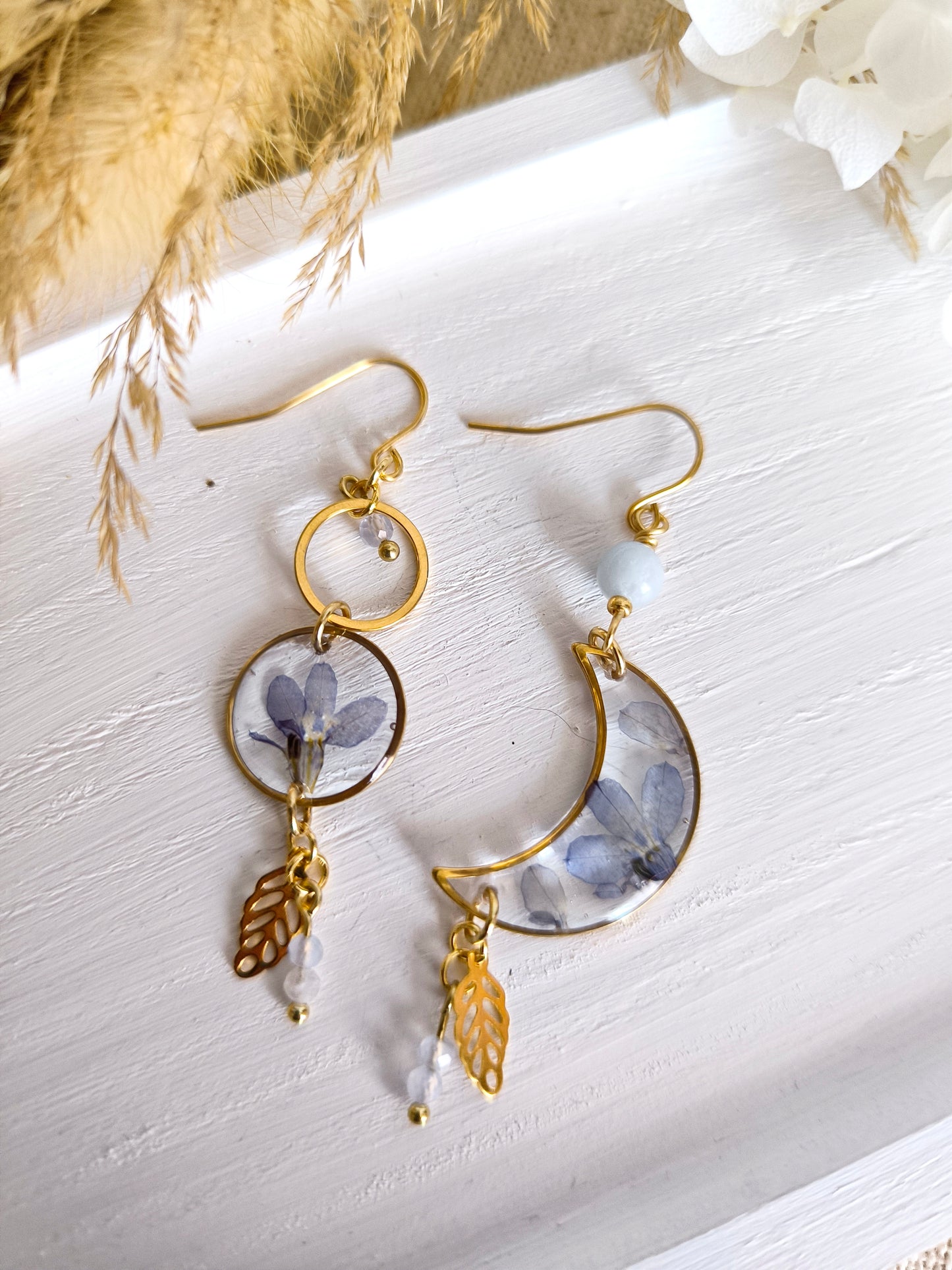 Boucles d'oreilles asymétriques en forme de lune et de soleil contenant une fleur séchée et des pierres fines.