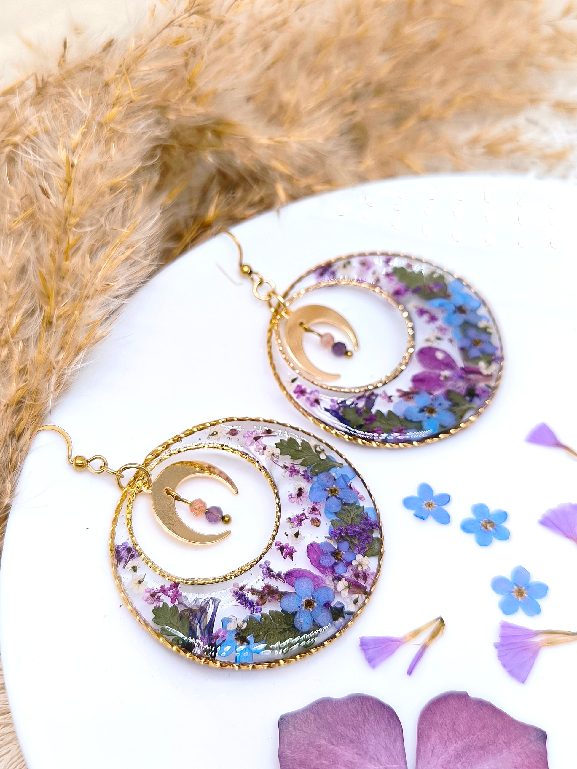 Boucles d'oreilles maxi créoles lunaires composées de fleurs séchées encapsulées dans une résine transparente.