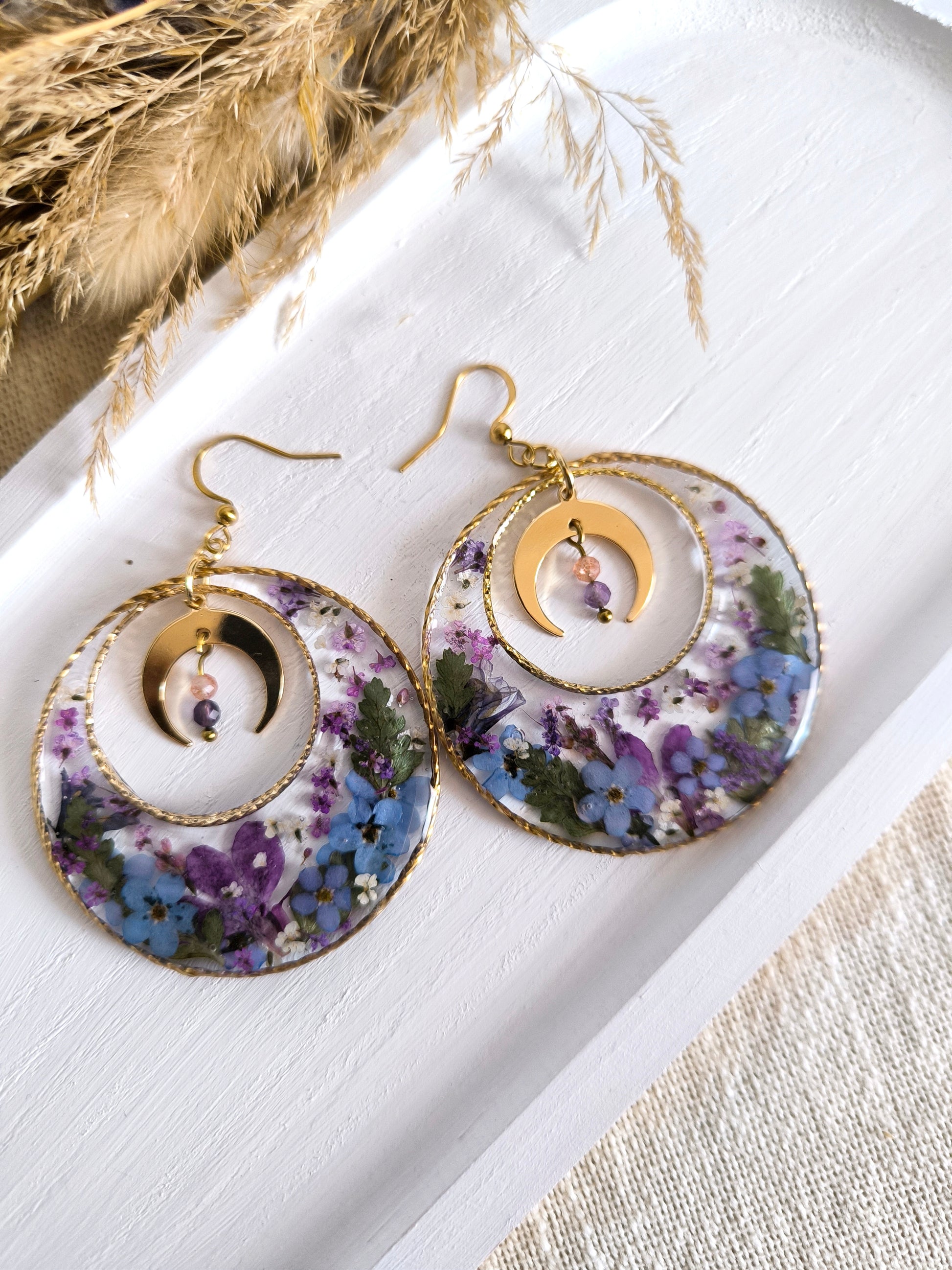 Boucles d'oreilles maxi créoles lunaires composées de fleurs séchées encapsulées dans une résine transparente.